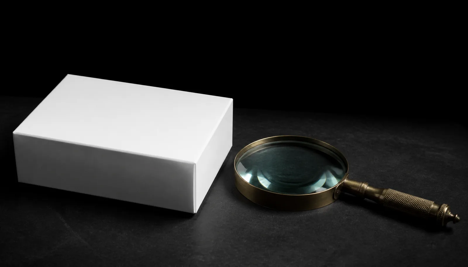 Caixa branca lisa fechada ao lado de uma lupa clássica sobre mesa escura, em luz dramática